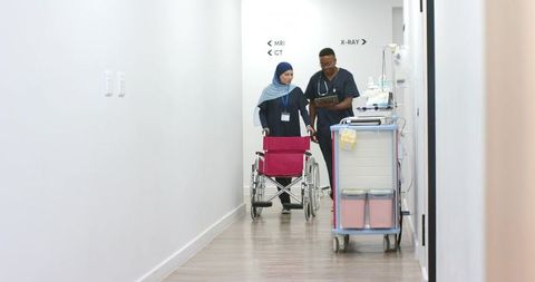 African american nurse and colleague walking hospital hallway pushing wheelchair consulting tablet