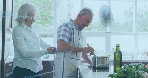 Senior couple cooking together in sunlit kitchen stirring pot while partner ties apron