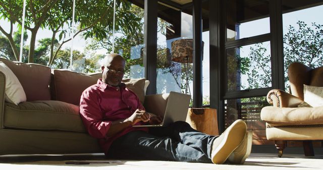 Man sitting on floor in living room, working on laptop with sunlight streaming in. Ideal for content related to remote work, home office setups, productivity, relaxation, or modern lifestyles.