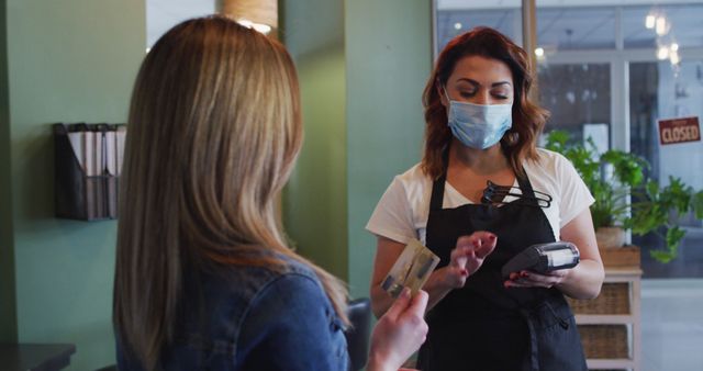 Waitress wearing mask holds card reader as customer hands over credit card. Suitable for illustrating pandemic dining protocols, contactless payment methods, and hospitality industry adaption during COVID-19.