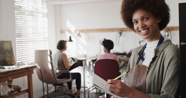 A woman with afro hair is smiling and holding a notebook in a creative workshop while other women work in the background. She wears an apron and is surrounded by artistic tools, suggesting an active and collaborative environment. This image can be used to showcase creative workspaces, artisanal craftsmanship, teamwork, organization, and planning. Ideal for promoting workshops, artisan businesses, creativity, or team-building activities.