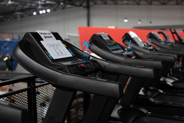 Treadmills in a gym with 'do not use' signs and hand sanitizer bottles placed on them for social distancing measures. Useful for illustrating health and safety protocols in fitness centers during the pandemic. Ideal for articles, blogs, and promotional materials related to gym safety, COVID-19 precautions, and public health guidelines.