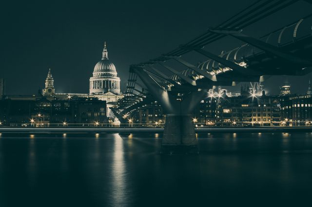 Nighttime image capturing illuminated St. Paul's Cathedral and Millennium Bridge reflecting over Thames River in London. Ideal for highlighting London landmarks, travel promotions, architectural studies, and urban lifestyle magazines. Use in content relating to nightlife, sightseeing, or European vacation planning.