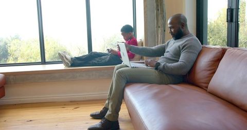 African american father working on laptop while son playing on smartphone at window seat