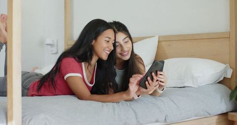 Diverse female friends sharing smartphone on cozy bed