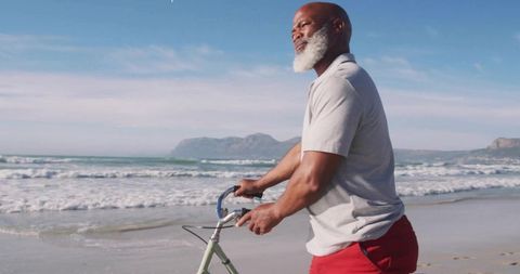 Senior man walking bicycle along sunlit beach with ocean waves and distant headlands