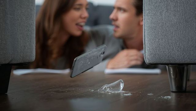 Smartphone falling and shattering on hardwood floor near sofa while couple watching