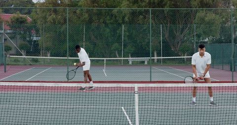 Diverse male tennis partners practicing doubles on outdoor hard court showing serving and teamwork