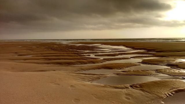 This photo captures a tranquil beach scene during low tide, with sand patterns and water reflections under a dramatic, cloudy sky. Suitable for promoting travel destinations, illustrating nature's beauty, or creating a relaxing ambiance in interior design.