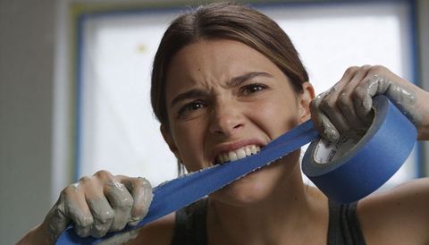 Determined woman biting blue painter tape while pulling roll during diy renovation, masked window