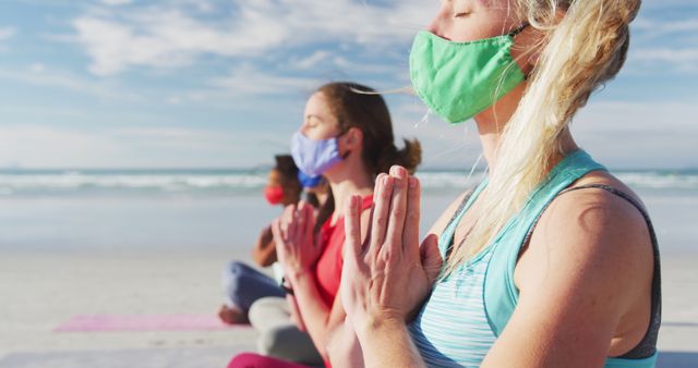 Group of diverse female friends wearing face masks meditating at the beach. healthy active lifestyle, outdoor fitness and wellbeing.