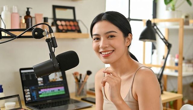 Asian beauty creator recording podcast with boom mic, laptop editing, smiling toward camera