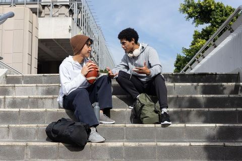 Diverse teen friends sitting on campus steps holding football and chatting between classes