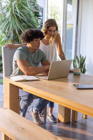 Young diverse couple working together on laptop at sunlit wooden dining table