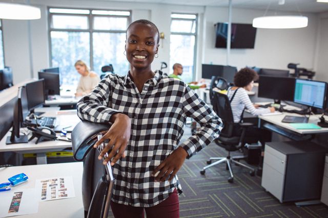 Confident African American businesswoman standing and smiling in a modern open-plan office with a diverse team working in the background. Ideal for illustrating team collaboration, workplace diversity, modern business environments, and professional confidence.