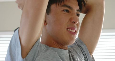 Young man raising arms showing armpits near window blinds wearing gray tee and earring