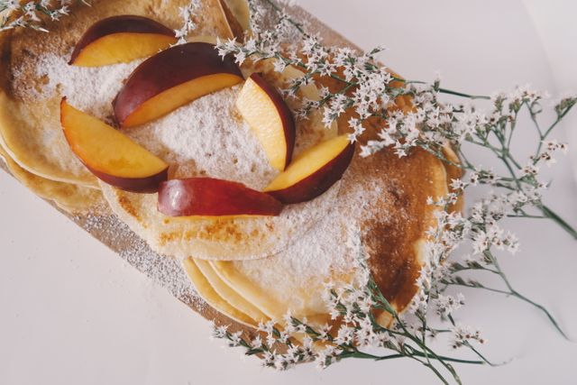 Pancakes with sliced peaches and delicate flower petals are topped with a dusting of powdered sugar. This visually appealing dish exudes freshness and sweetness, making it ideal for use in food blogs, culinary magazines, recipe websites, or restaurant menus focusing on brunch or breakfast items.