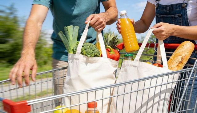 Couple carrying reusable tote bags filled with fresh produce, bread and juice in cart