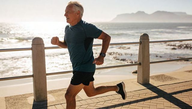 Active senior man jogging along sunlit seaside promenade wearing smartwatch and shorts