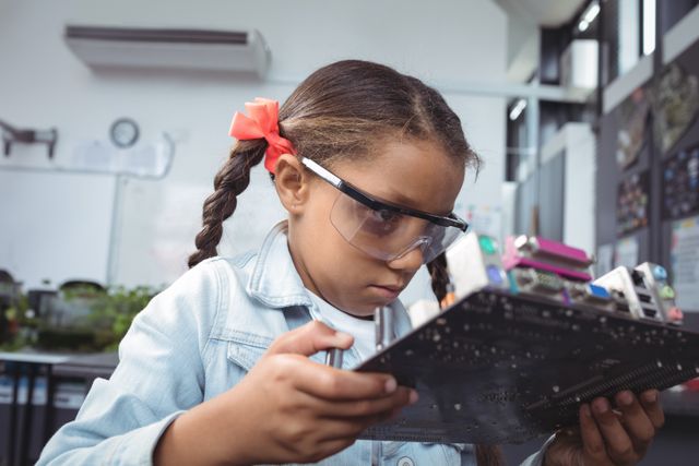Young girl in an electronics lab carefully examining a circuit board, wearing safety glasses. Ideal for use in educational materials, STEM program promotions, technology and science learning resources, and articles about young innovators and future scientists.