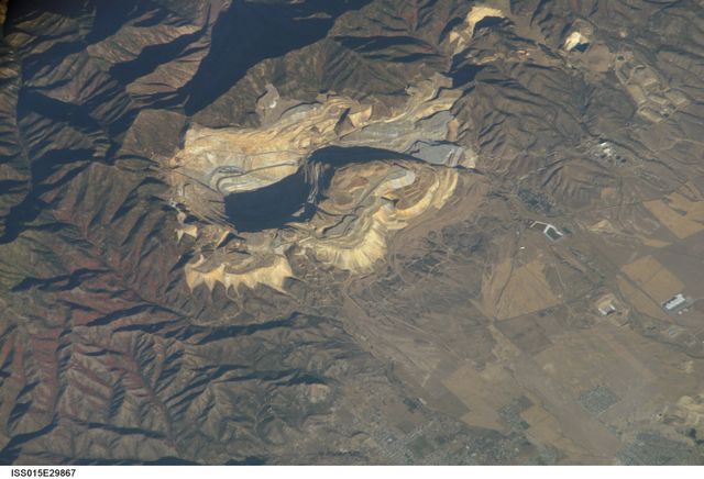 ISS015-E-29867 (20 Sept. 2007) --- Bingham Canyon Mine, Utah is featured in this image photographed by an Expedition 15 crewmember on the International Space Station. The Bingham Canyon Mine (center) located approximately 32 kilometers to the southeast of Salt Lake City, UT is one of the largest open-pit mines in the world, measuring over 4 kilometers wide and 1,200 meters deep. The mine exploits a porphyry copper, a type of geological structure formed by crystal-rich magma moving upwards through pre-existing rock layers. As the magma cools and crystallizes (forming an igneous rock with large crystals in a fine-grained matrix, known as a porphyry), hot fluids circulate through the magma and surrounding rocks via fractures. This process of hydrothermal alteration typically forms copper-bearing and other minerals in spatial patterns that a geologist recognizes as a potential porphyry copper deposit. Parallel benches (stepped terraces), visible along the western pit face (center left), range from 16 to 25 meters high - these provide access for equipment to work the rock face, as well as maintaining stability of the sloping pit walls. A dark, larger roadway is also visible directly below the benches. Brown to gray, flat topped hills of gangue (waste rock) surround the pit, and are thrown into sharp relief by shadows and the oblique viewing angle of this image. Leachate reservoirs associated with ore processing are visible to the south of the city of Bingham Canyon, UT (right).