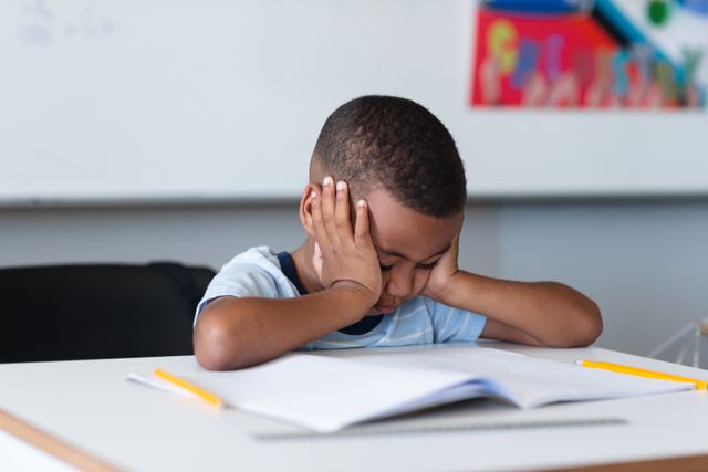 Young African American boy sitting at a desk in a classroom with his head in his hands, concentrating on his schoolwork. Ideal for use in educational materials, school websites, and articles about childhood education, learning challenges, and student concentration.