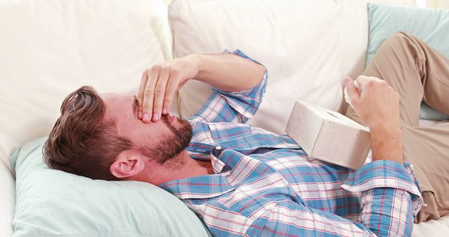 A middle-aged Caucasian man appears unwell as he lies on a couch with a book, holding his forehead, indicating a headache or fever. His expression and posture suggest discomfort or illness, emphasizing the need for rest or medical attention.