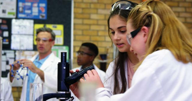 Caucasian teenage girls in lab coats are examining a sample through a microscope in a science class, with copy space. A diverse group of students and a teacher in the background suggest an interactive learning environment.