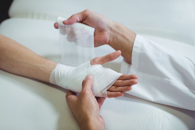 This image shows a physiotherapist carefully bandaging an injured hand of a patient in a clinical setting. It is ideal for use in healthcare, medical care, and rehabilitation contexts, illustrating professional treatment and patient care. Suitable for websites, brochures, and educational materials related to medical services and injury recovery.