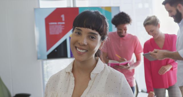 Businesswoman stands confidently in a modern office setting with a smile, while team members collaborate in background. Ideal for use in corporate materials, presentations, and articles focused on teamwork, leadership, and modern work environments.