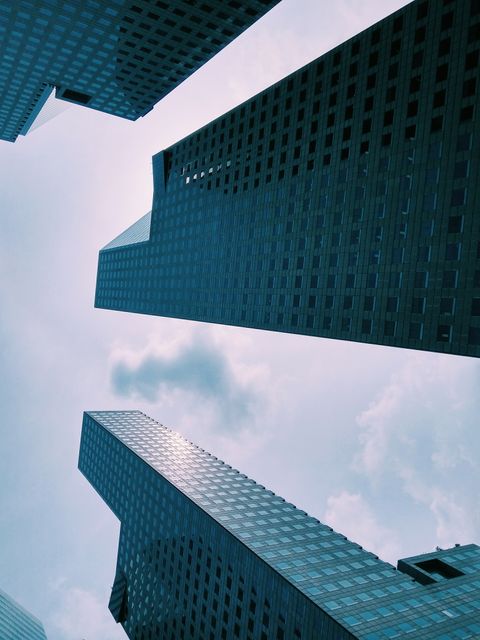 This image features a low angle view of modern skyscrapers, emphasizing their height against a cloudy sky. It showcases the urban landscape with high-rise buildings and office spaces. Ideal for use in projects related to city planning, corporate publications, real estate, or urban development. The dramatic perspective captures the essence of a bustling city skyline.