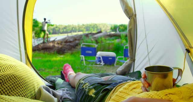 This image shows a person relaxing inside a tent, holding a metal mug and looking out at the lush outdoors. Visible are green trees, camping chairs, a cooler, and another person in the background. Ideal for promoting camping equipment, travel blogs, nature retreats, and outdoor adventure activities.
