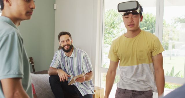 Three friends spending time together, one using a VR headset while the others watch and smile. They appear relaxed and comfortable in a modern living room, showcasing camaraderie and new technology. Useful for depicting concepts like social leisure, entertainment, innovation, and VR technology in a positive light.