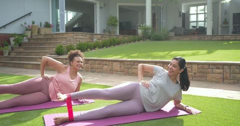 Diverse women exercising with resistance band doing side-lying leg lifts on backyard mats