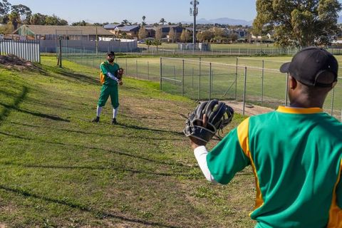 Baseball teammates warming up on sunny grassy hillside near fenced neighborhood ballpark