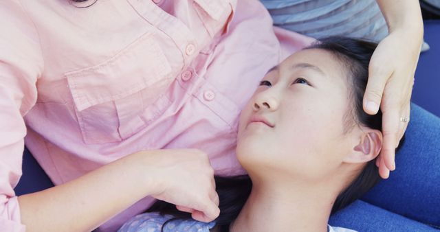 A young girl is lying on her mother's lap looking up at her. The mother gently cradles her daughter's face and head, showing comfort and affection. Both are in casual clothing, suggesting a relaxed setting. This image is suitable for use in parenting articles, family care topics, maternal affection highlights, or emotional well-being promotions.