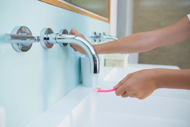 Child holding toothbrush under running water at bathroom sink. Ideal for topics on dental hygiene, morning routines, children's health, and home care. Useful for educational materials, health campaigns, and parenting blogs.