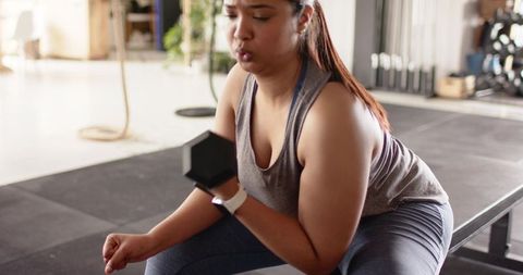 Woman lifting hex dumbbell while sitting on bench in gym, strength training focus