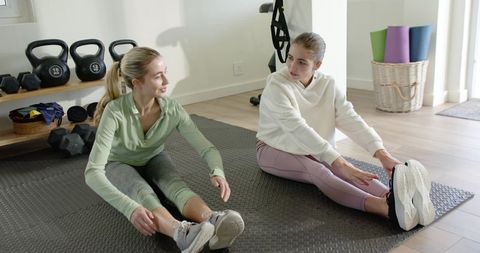 Two women stretching on home gym mat with kettlebells and yoga mats, sunlit wellness