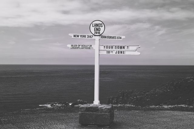 Vintage signpost at Land's End, indicating distances to New York, John O'Groats, Isles of Scilly, and Longships Lighthouse against an ocean view. Useful for travel magazines, tourism advertisements, and landmark illustrations.