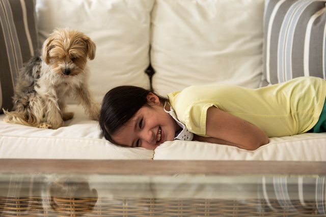 Little caucasian girl lying on the couch at home smiling beside her dog. their reflection can be seen on the table in the foreground.