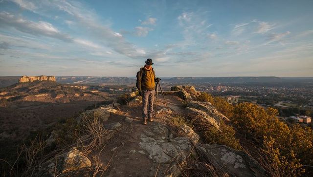 Photographer adjusting camera on tripod atop rocky ridge overlooking valley during golden hour