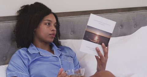 African american woman relaxing in satin pajamas reading paperback in cozy bed, serene