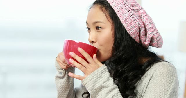 Young woman drinking from red mug while wearing knit hat indoors. Suitable for themes around winter, relaxation, comfort, leisure activities, casual lifestyle, and cozy moments. Can be used in lifestyle blogs, winter fashion articles, or advertisements for warm beverages.