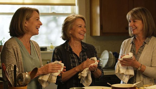 Three women laughing and drying dishes together in cozy sunlit home kitchen