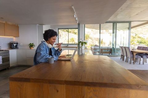 African american man working from home on laptop at modern kitchen island with terrace