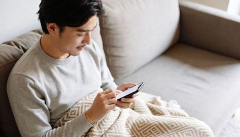 Asian man relaxing on sofa scrolling smartphone under cozy blanket at home