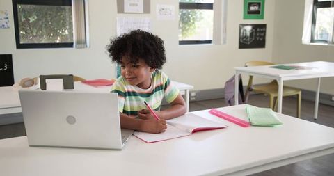 African American boy studying with laptop and notebook in modern classroom