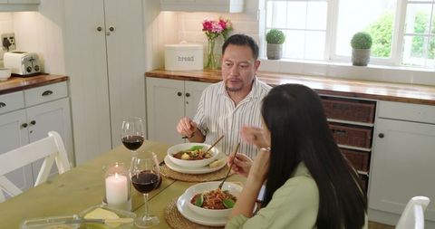 Asian couple enjoying cozy rustic kitchen dinner with noodles, wine, candlelight, sunlight