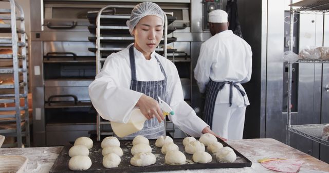 Professional baker spraying water on dough buns in a commercial kitchen, showing a step in bread making process. Ideal for illustrating culinary techniques, food industry environments, professional baking, teamwork in kitchens, and daily activities of chefs.